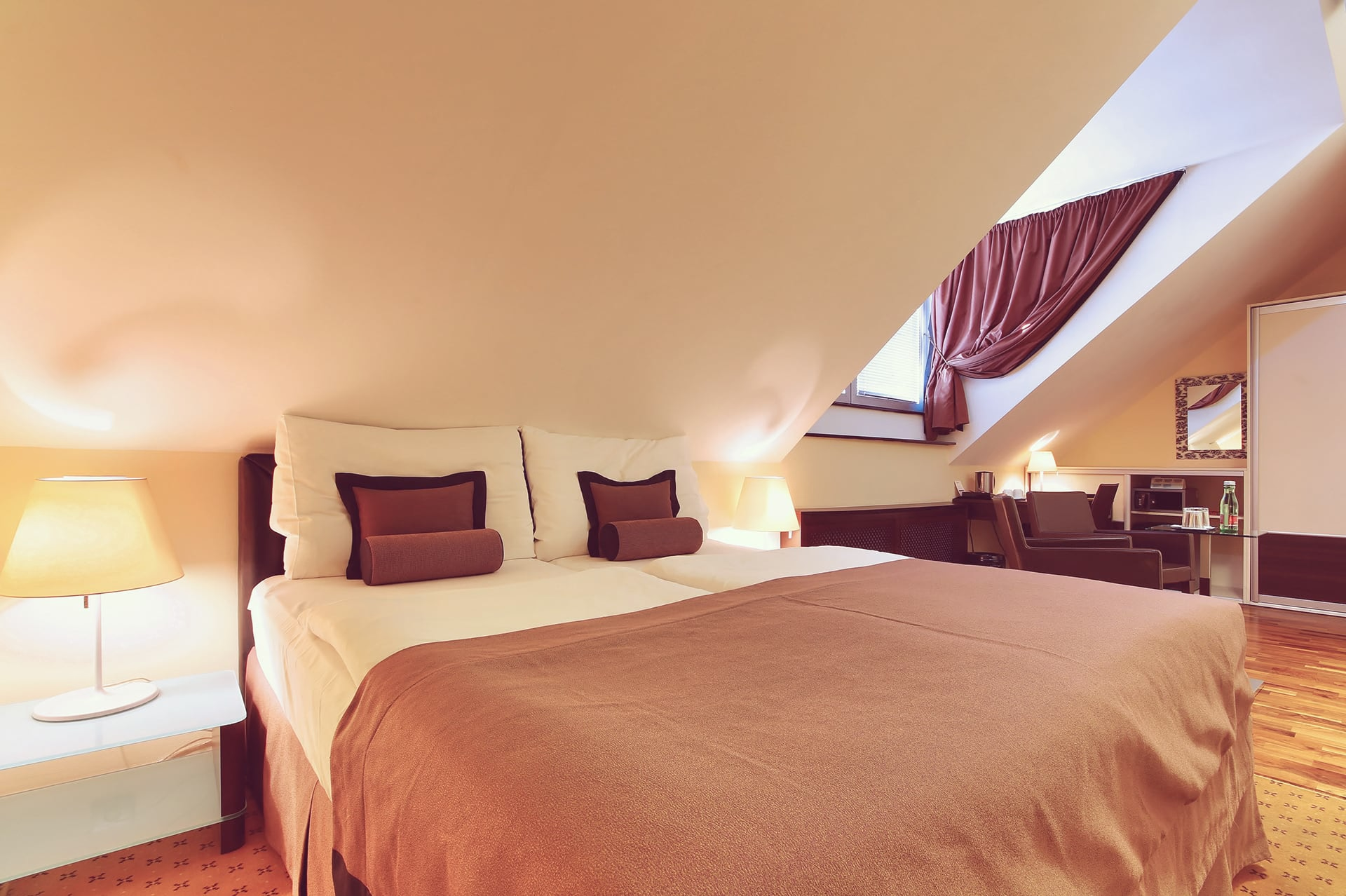 Angle shot of double bed under sloped ceiling with bedside lamps in an attic room at Skaritz Hotel.
