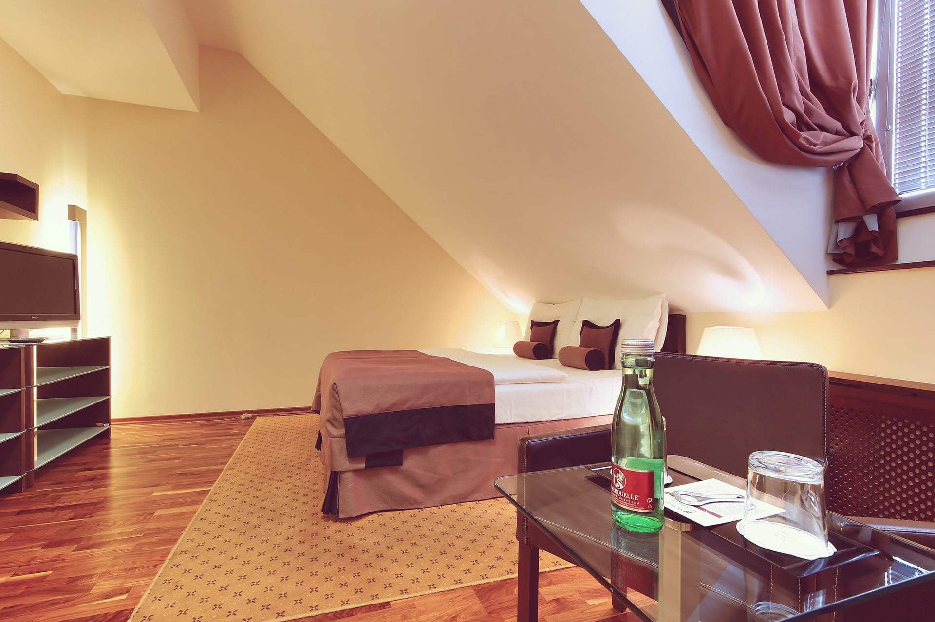 Sloped ceiling attic room with double bed, TV console, and glass coffee table with mineral water.