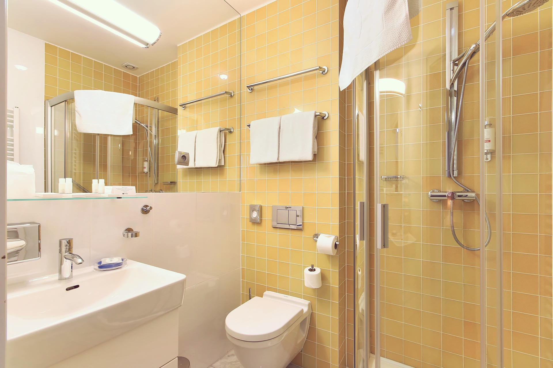 Modern hotel bathroom with warm yellow tiles, glass shower stall, wall-hung toilet, and vanity sink.