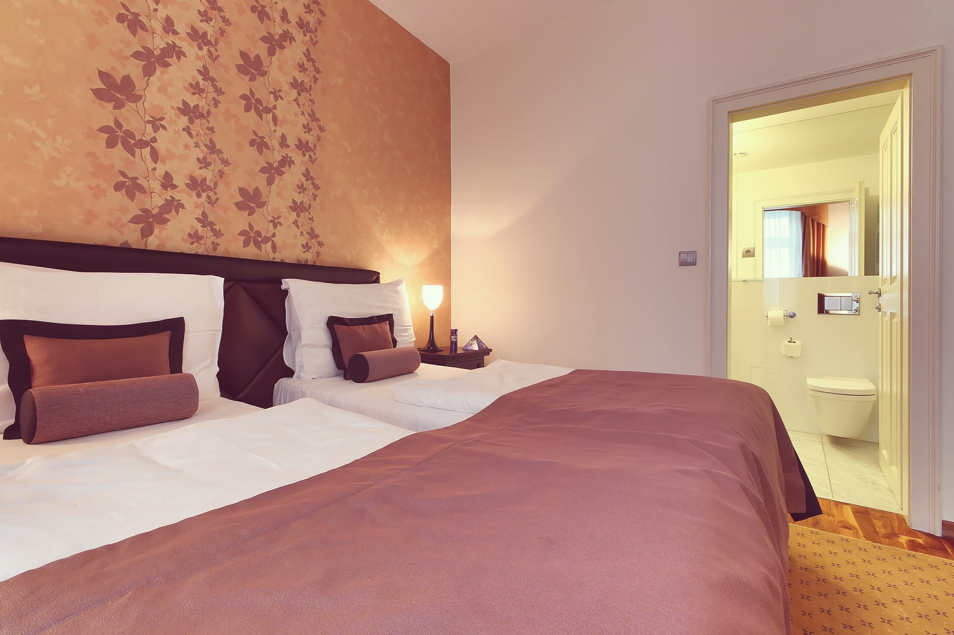 Double room bed with brown throw blanket and open door to the en-suite bathroom at Skaritz Hotel.