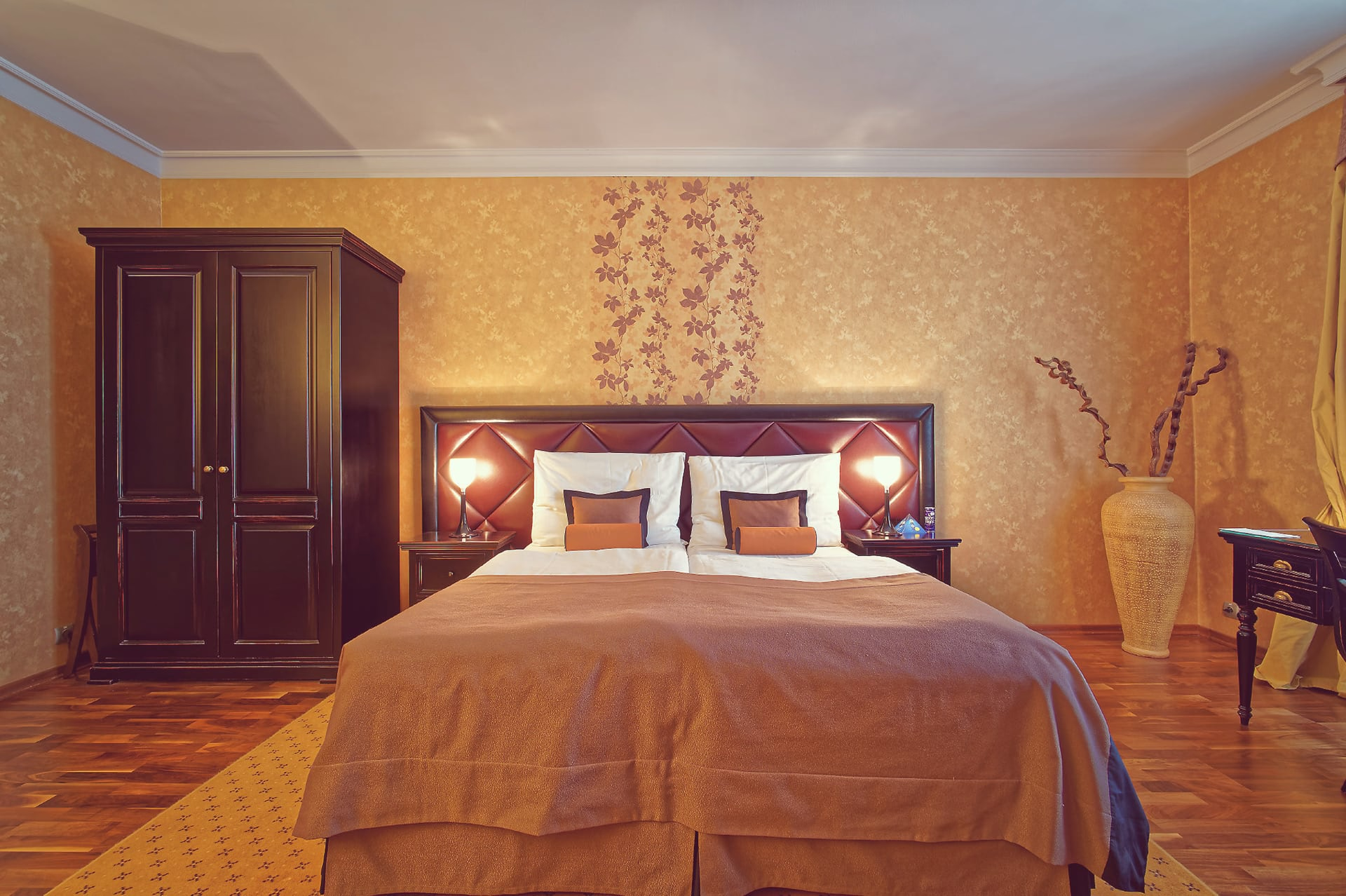 Front view of king-size bed with nightstands, dark wood wardrobe, vase and decorative wall pattern at Skaritz Hotel.