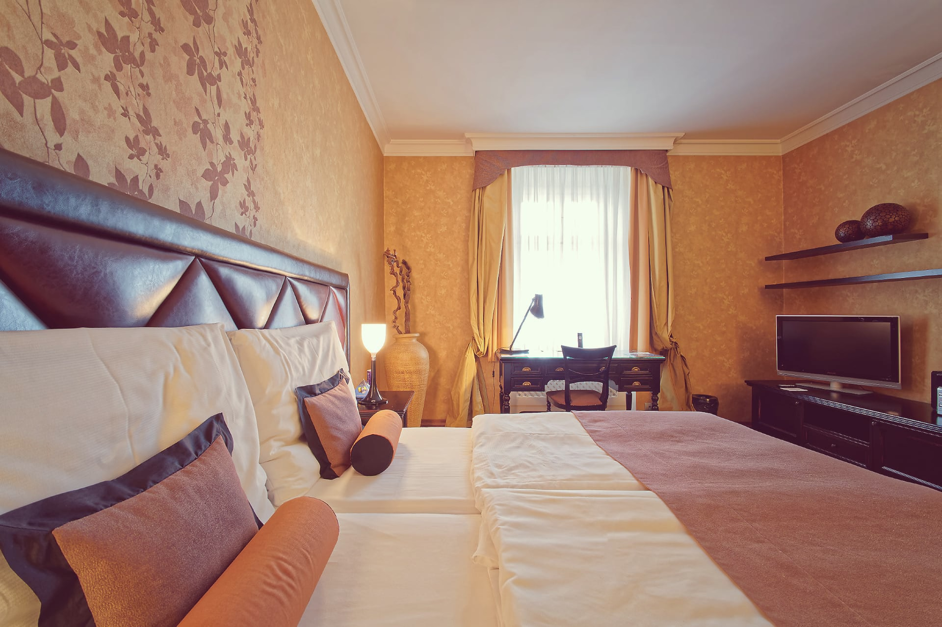 Close-up of bed pillows with writing desk by the window and TV unit in the background.