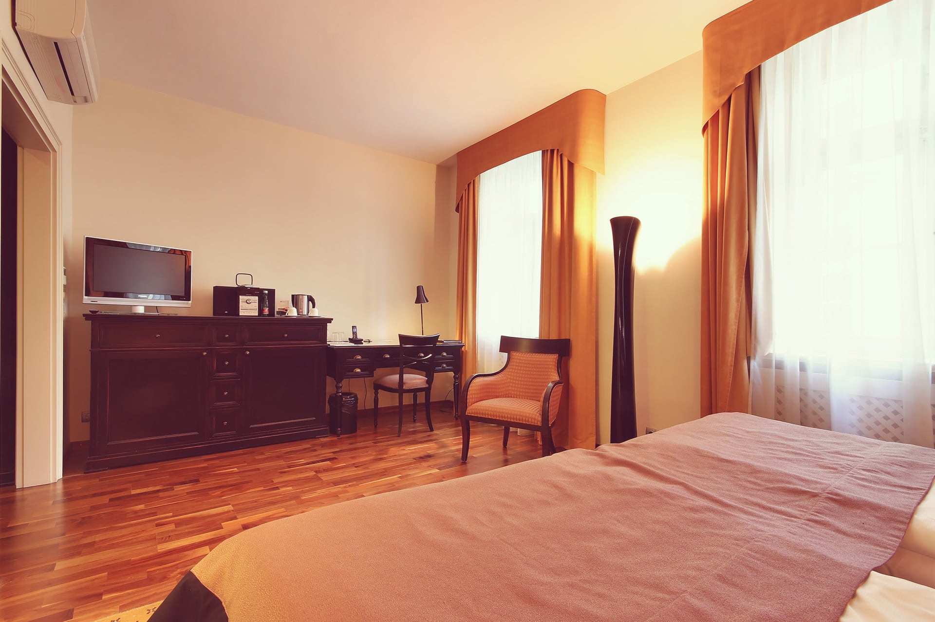 Hotel room interior showing dark wooden TV dresser, work desk, and armchair next to windows.
