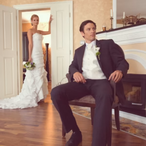 Elegantly dressed groom sitting near a fireplace with a bride in a white gown standing in the doorway during a wedding photo shoot in a Skaritz Hotel suite.